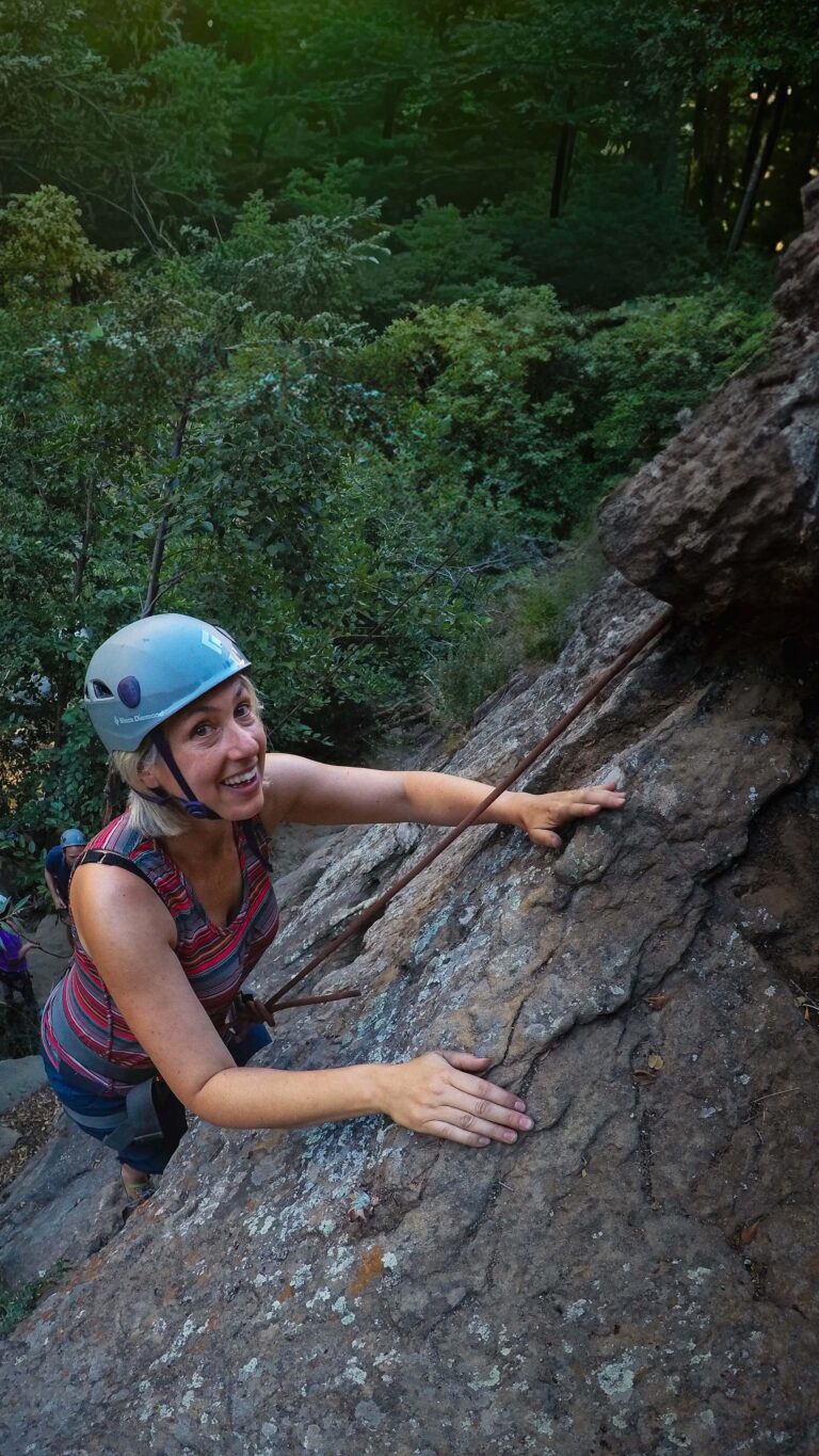 Female climber smiling while scaling a textured sandstone face surrounded by lush green foliage in Santa Barbara.