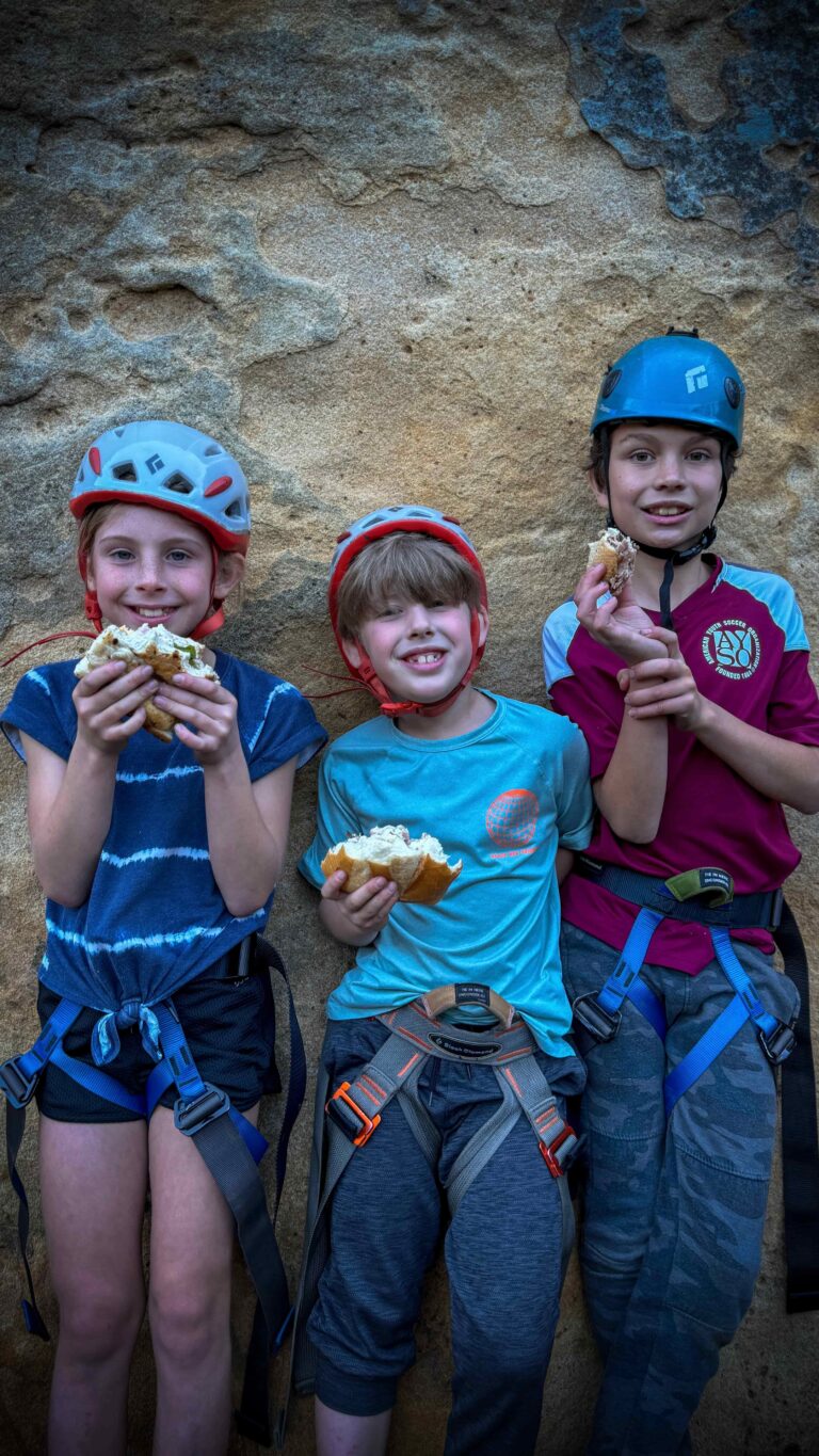 Three kids wearing climbing helmets and harnesses eating snacks during a guided Group Climbing Experience in Santa Barbara, standing against a sandstone wall.