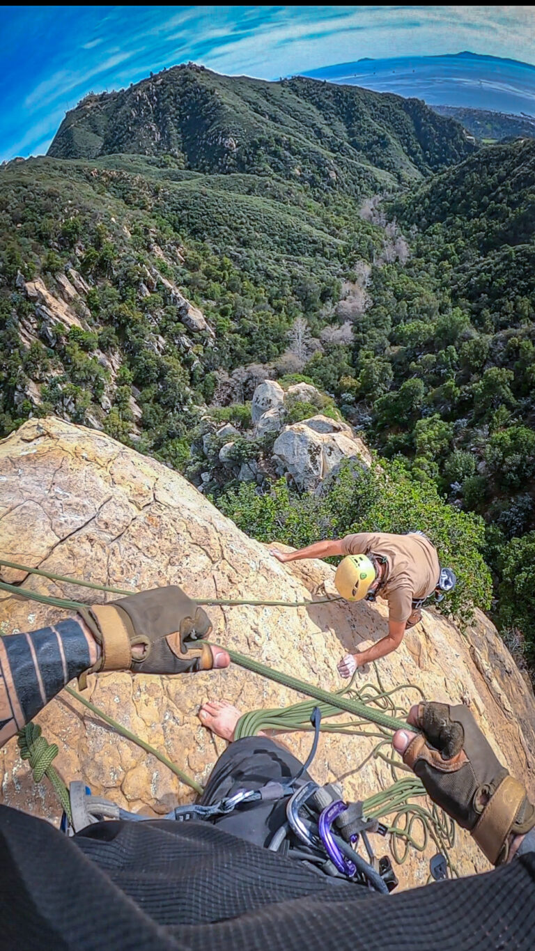 Belayer at the summit managing ropes as a climber tops out on a multipitch climb with expansive Santa Barbara mountain and coastline views.