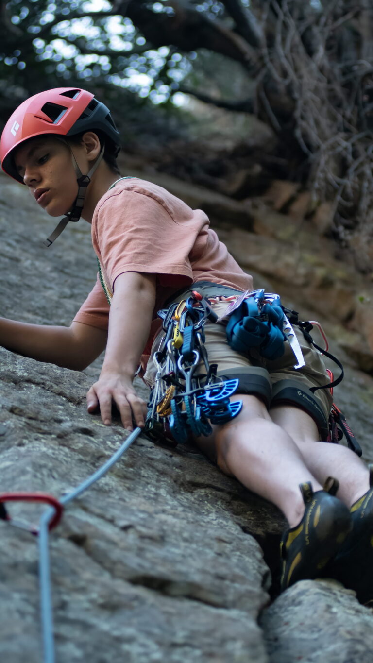 Young climber wearing a helmet and carrying a full rack of climbing gear while ascending a rock face during a lesson.